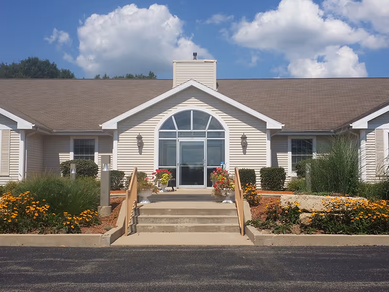 Front exterior view of a single-story building with beige siding and a brown roof. There is a central entrance with glass double doors topped by a large arched window. Concrete steps with handrails lead up to the entrance, flanked by flower beds with yellow and red flowers and green shrubs. The sky is blue with some white clouds.