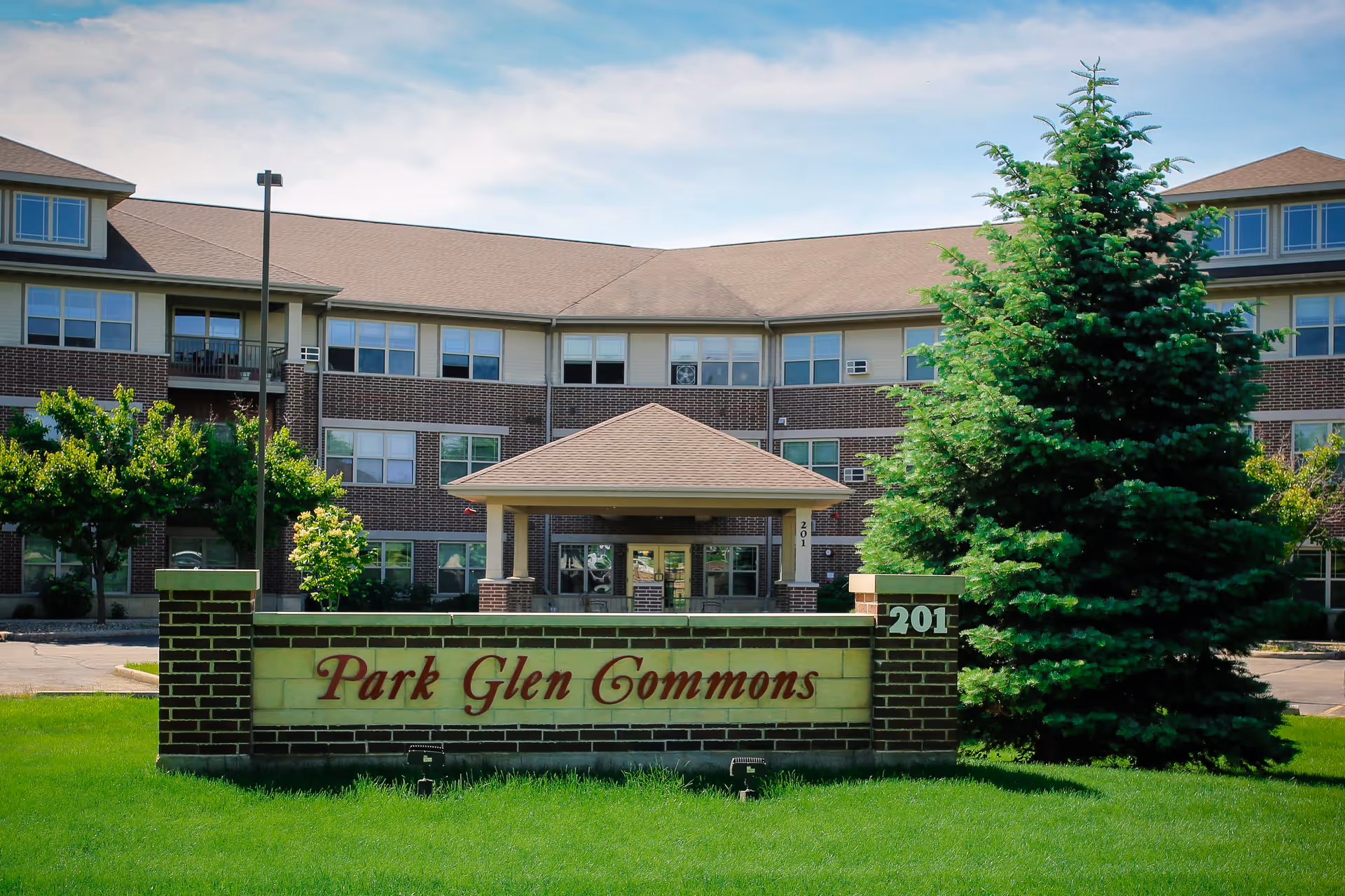 Exterior view of Park Glen Commons, a multi-story brick building with numerous windows and a covered entrance. In front of the building is a brick sign with the name 'Park Glen Commons' and the number 201. There are green trees and a well-maintained lawn surrounding the sign and building.