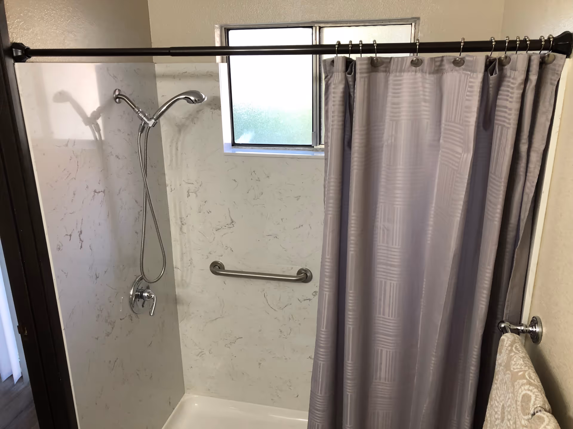 A shower area with a silver handheld showerhead attached to a wall with a marble-like pattern. There is a silver grab bar mounted on the wall and a small frosted window above it. A gray shower curtain hangs from a black shower rod, and a beige patterned towel is hanging on a silver towel ring on the right side.