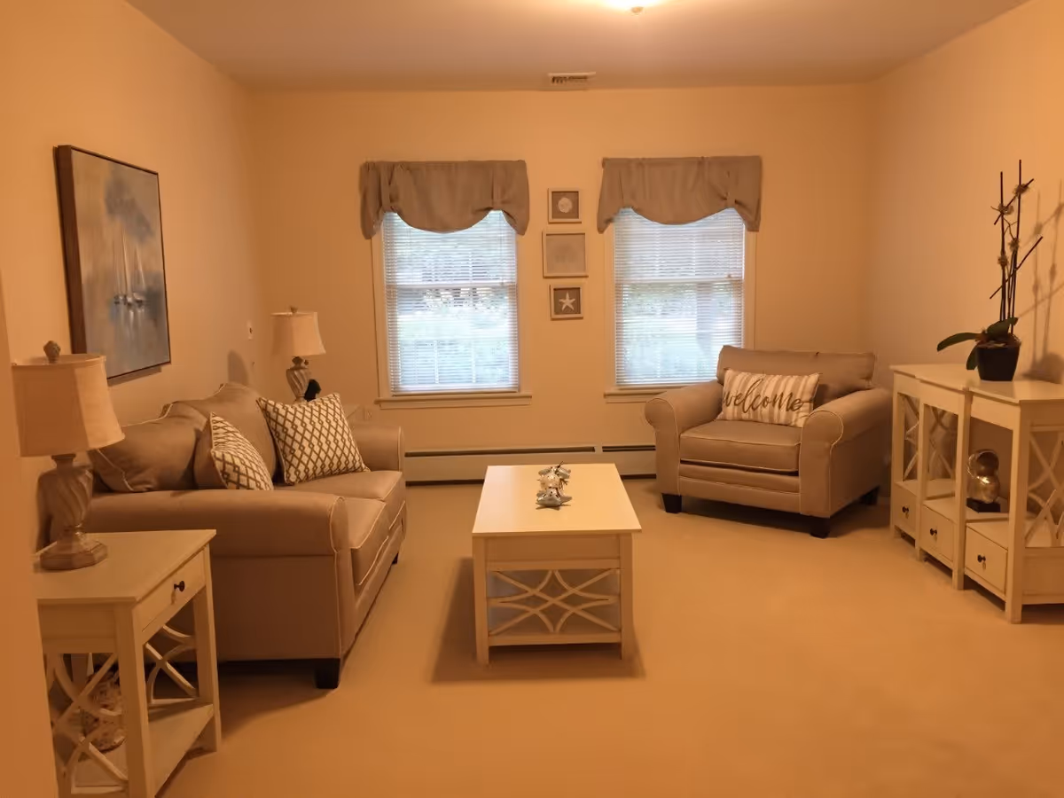 A cozy living room with beige walls and carpet. The room features a beige sofa with patterned pillows on the left, a matching armchair with a 'welcome' pillow on the right, and a white coffee table in the center. There are two windows with beige valances and blinds on the back wall, framed by three small decorative pictures. On the left side, there is a side table with a lamp and a large painting above the sofa. On the right side, there is a white cabinet with decorative items and a potted plant on top.