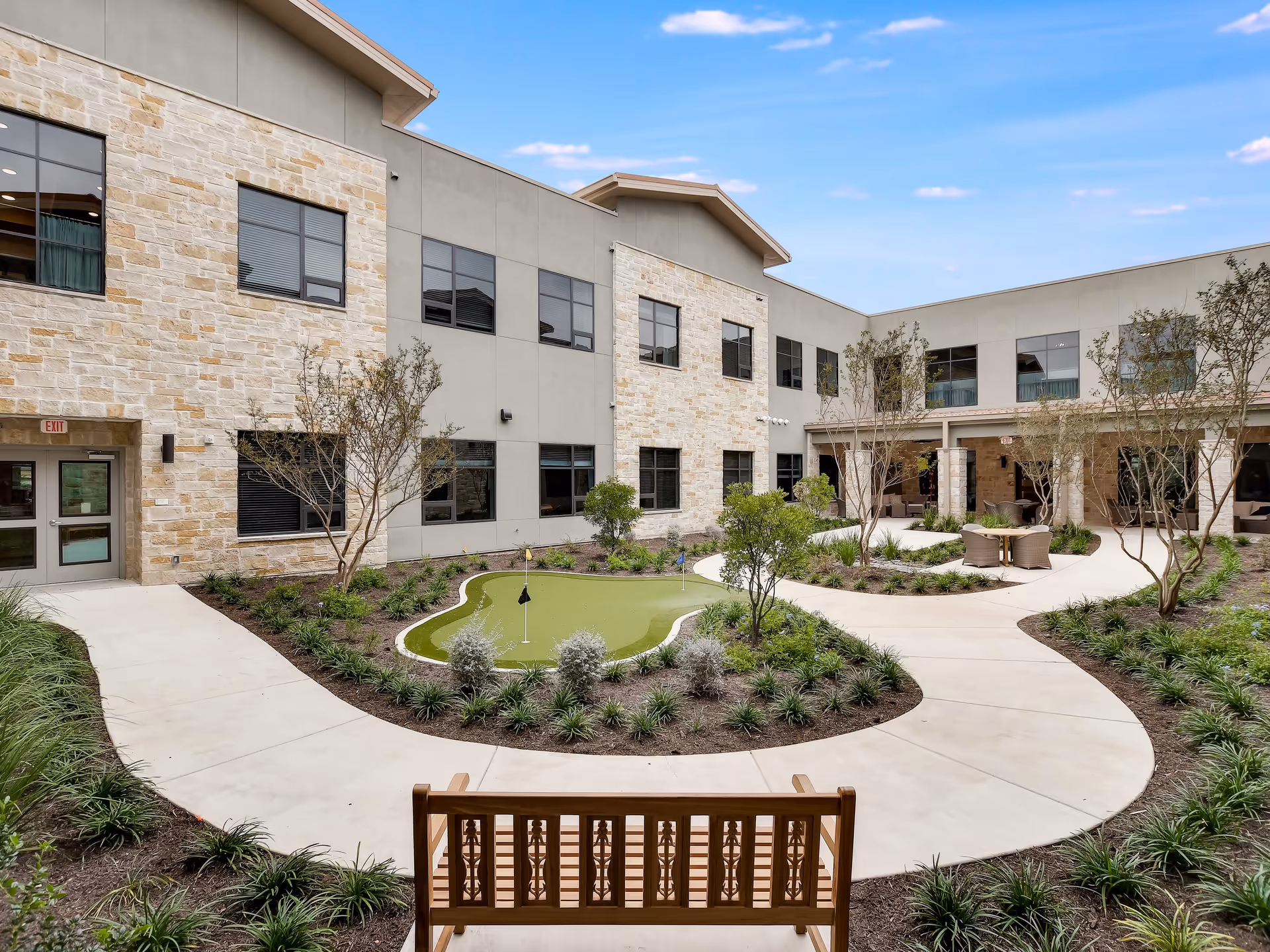 A landscaped interior courtyard with winding walkways, a small putting green, benches and seating surrounded by a two-story building.
