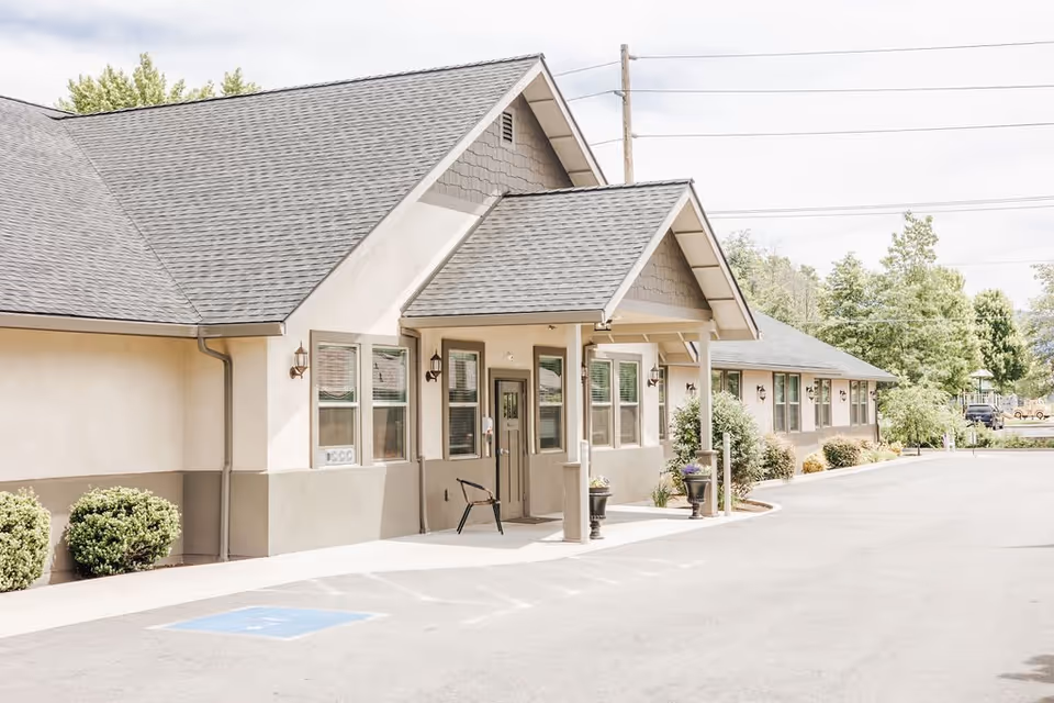 Exterior view of a single-story building with a gray shingled roof and beige walls, featuring a covered entrance with a chair and potted plants near the door. The building is surrounded by small bushes and trees, with a paved driveway and a handicap parking space visible in front.