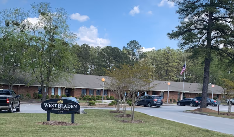 Exterior view of West Bladen Assisted Living And Memory Care facility showing a single-story brick building with a parking lot in front, several cars parked, trees, and a sign on the grass that reads 'West Bladen Assisted Living 714'. The sky is blue with some clouds.