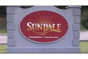 A large stone sign with a red oval center displaying the name 'Sundale' in large letters, with the words 'Rehabilitation | Long Term Care' below it. The sign is situated outdoors with grass and a road visible in the background.