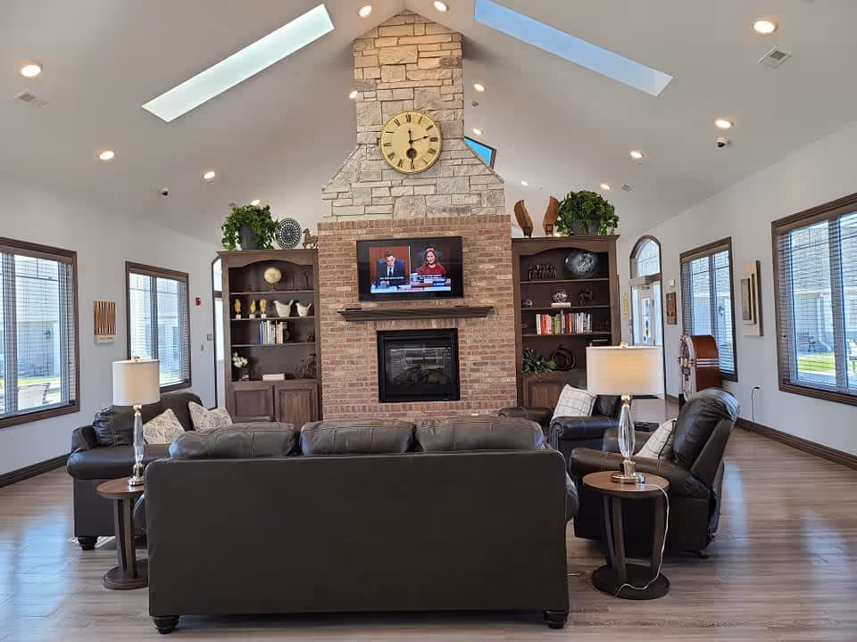 A spacious living room with a high vaulted ceiling featuring skylights and recessed lighting. The room has a large stone and brick fireplace with a clock above it and a flat-screen TV mounted on the brick. There are built-in wooden shelves on either side of the fireplace decorated with books, plants, and ornaments. The seating area includes dark leather sofas and armchairs arranged around the fireplace, with side tables and lamps. Large windows with wooden frames allow natural light to fill the room.