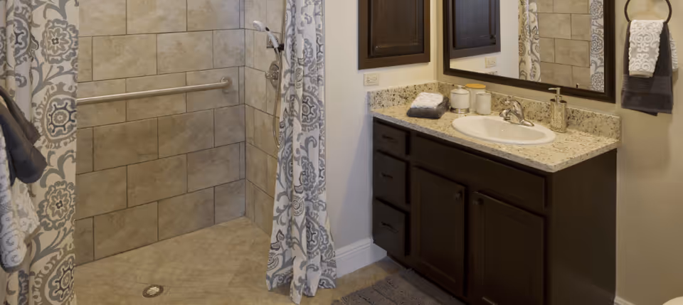 A bathroom featuring a tiled walk-in shower with a patterned shower curtain and a metal grab bar. Adjacent to the shower is a dark wood vanity with a granite countertop, a white sink, a mirror above, and neatly folded towels and containers on the counter. A towel ring with a white towel is mounted on the wall next to the vanity.
