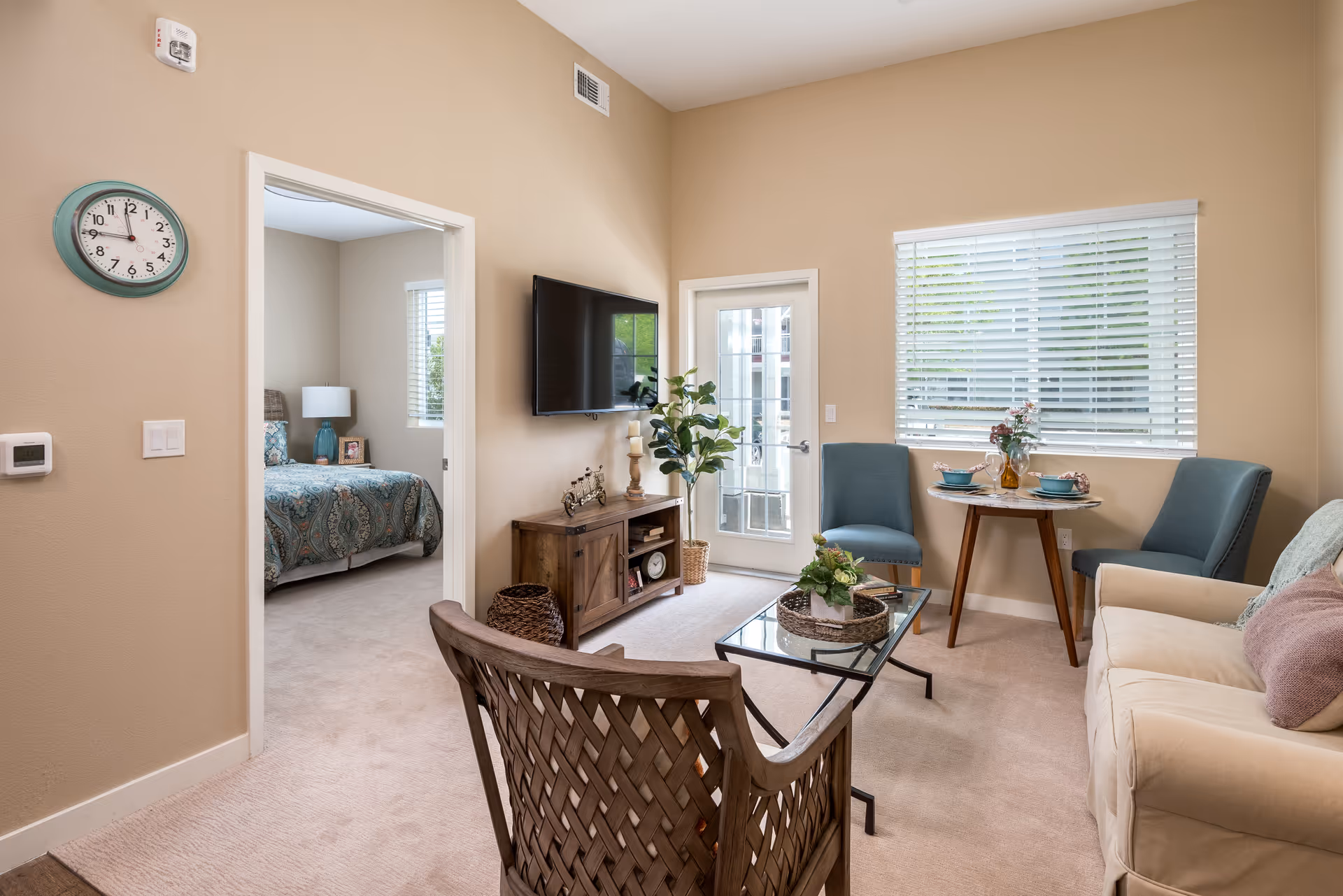 A cozy living room area with a beige sofa, two blue chairs, a small round dining table set for two, a glass coffee table with a plant centerpiece, and a wooden chair with a woven back. A flat-screen TV is mounted on the wall above a wooden cabinet. There is a door with glass panels and a window with white blinds letting in natural light. An adjacent bedroom with a bed covered in a patterned blue and green bedspread is visible through an open doorway.