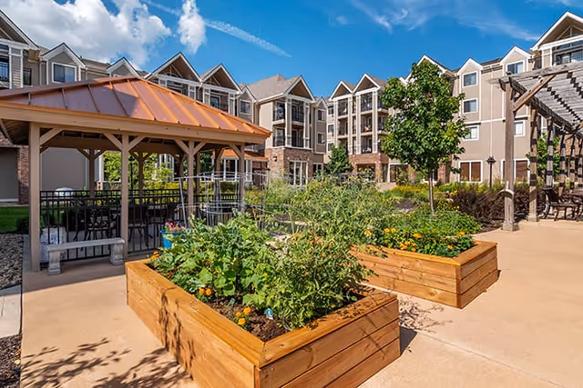 Outdoor courtyard area at Brookdale Wornall Place featuring raised wooden garden beds with plants, a gazebo with seating underneath, a pergola with chairs, and a multi-story residential building in the background under a blue sky with some clouds.