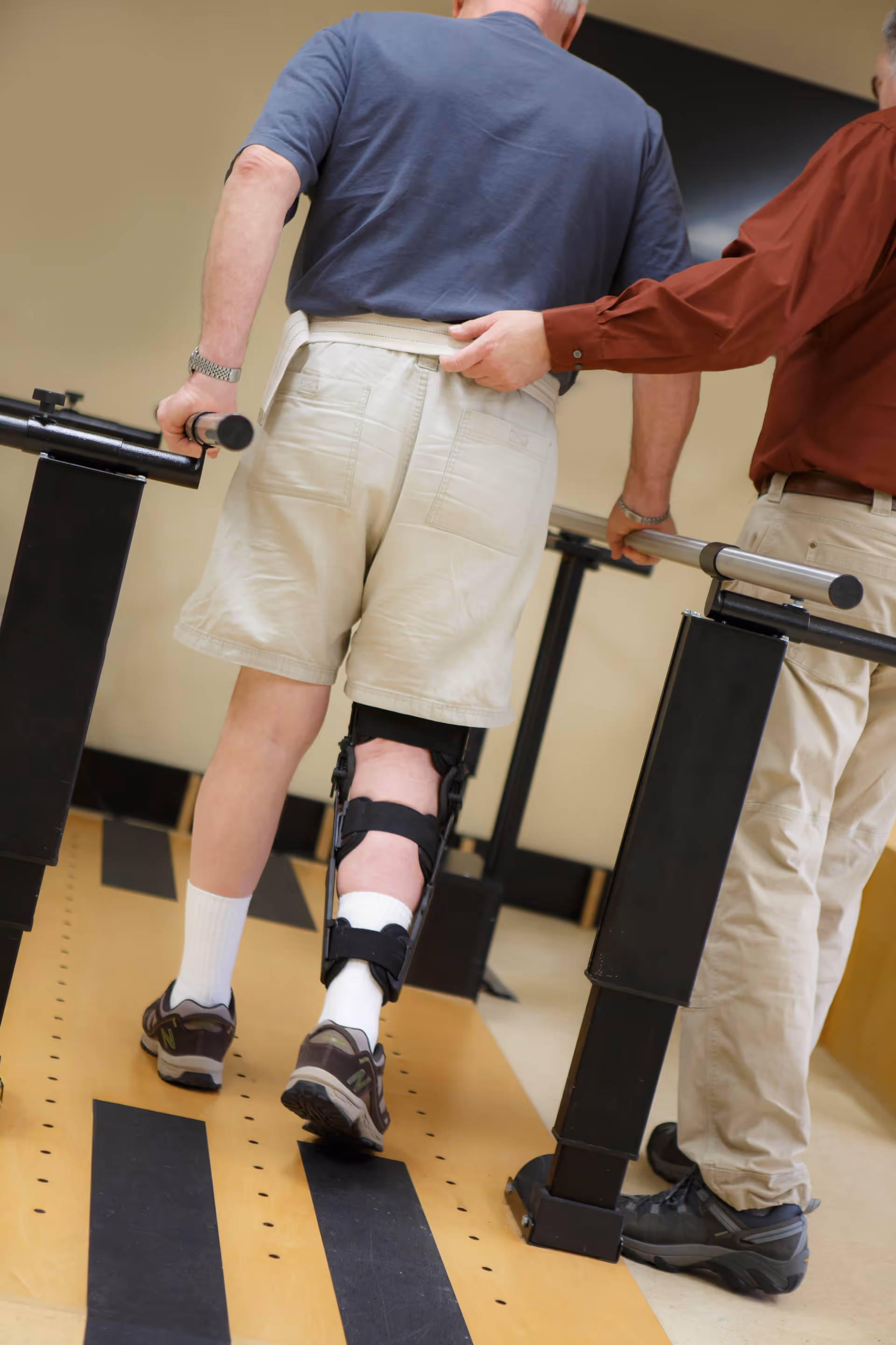 A person wearing a leg brace and shorts is walking with the support of parallel bars, assisted by another person holding their waist for support during physical therapy or rehabilitation.