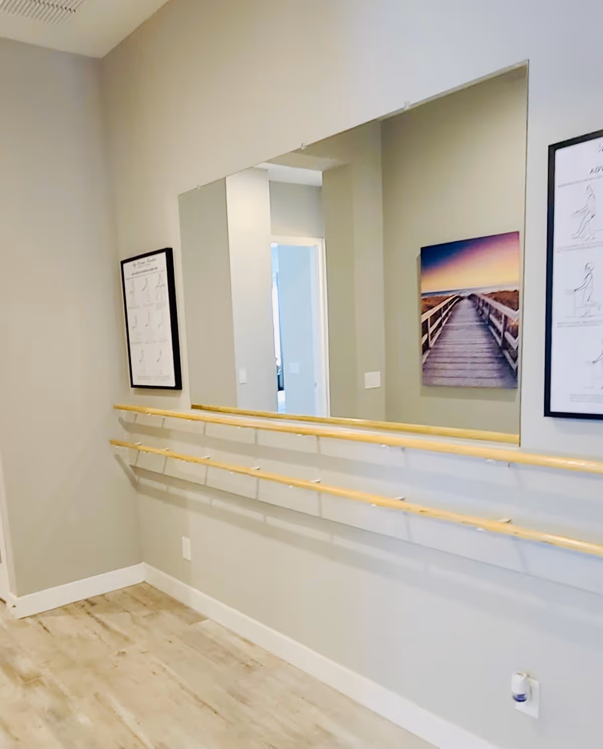 Interior room with light-colored walls and wooden flooring featuring two parallel wooden handrails mounted on the wall beneath a large mirror. The wall also has framed pictures, including one of a boardwalk leading to a sunset and another with exercise instructions.