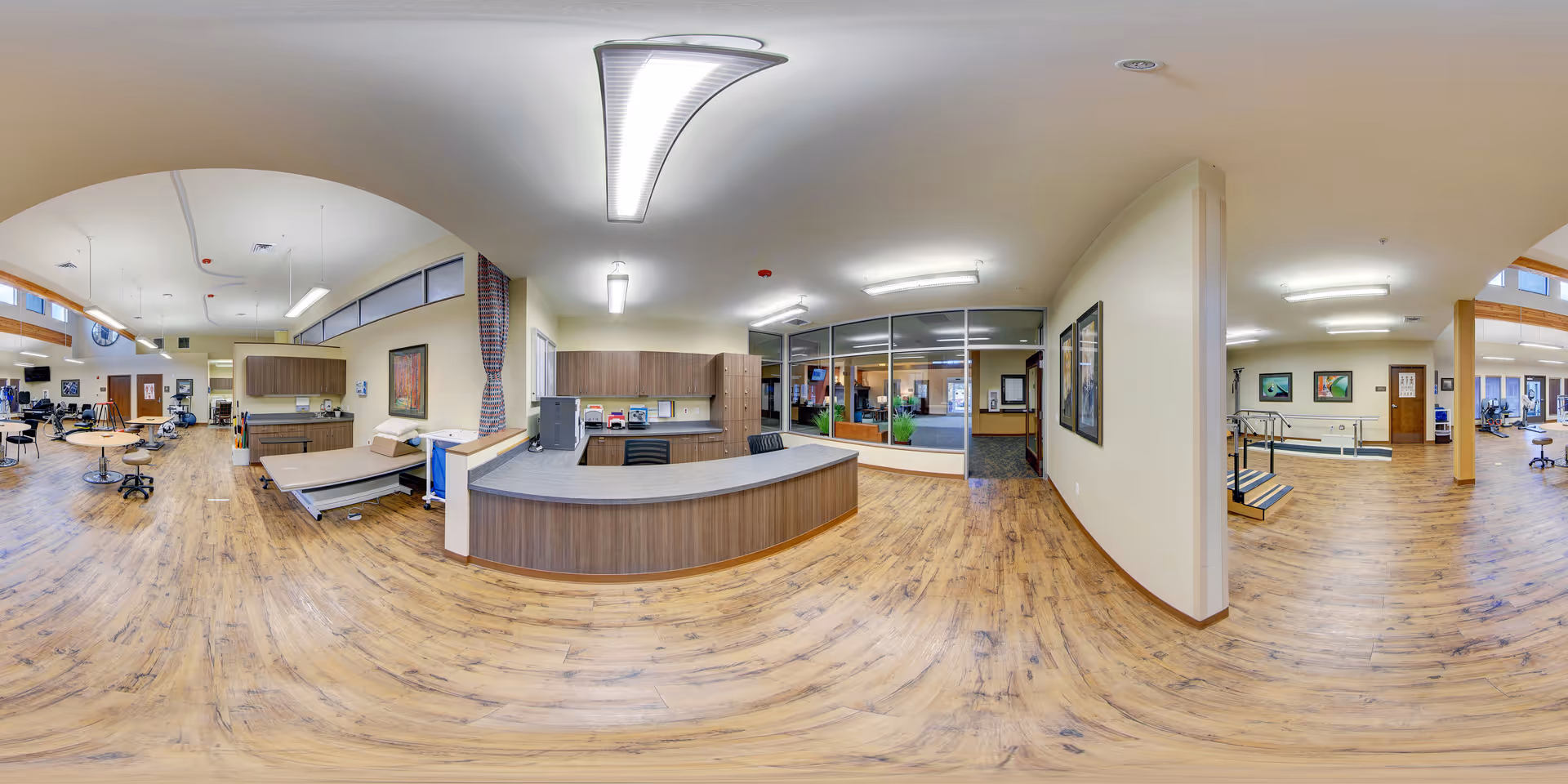 A spacious interior room with wood flooring and neutral-colored walls, featuring a reception desk with chairs, medical examination tables, exercise equipment, and framed artwork on the walls. The room is well-lit with ceiling lights and has large windows and glass partitions separating different areas.