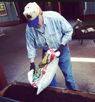 An elderly man wearing a light blue patterned shirt, blue jeans, black gloves, and a beige baseball cap is pouring soil from a bag into a raised garden bed outdoors. There is a folding chair and building structure in the background.