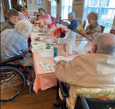 A group of elderly individuals seated around a long table engaged in arts and crafts activities. The table is covered with a pink tablecloth and various craft supplies, including paint bottles, paper cutouts, and markers. The room is well-lit with large windows and decorated with framed pictures and flowers on the table.