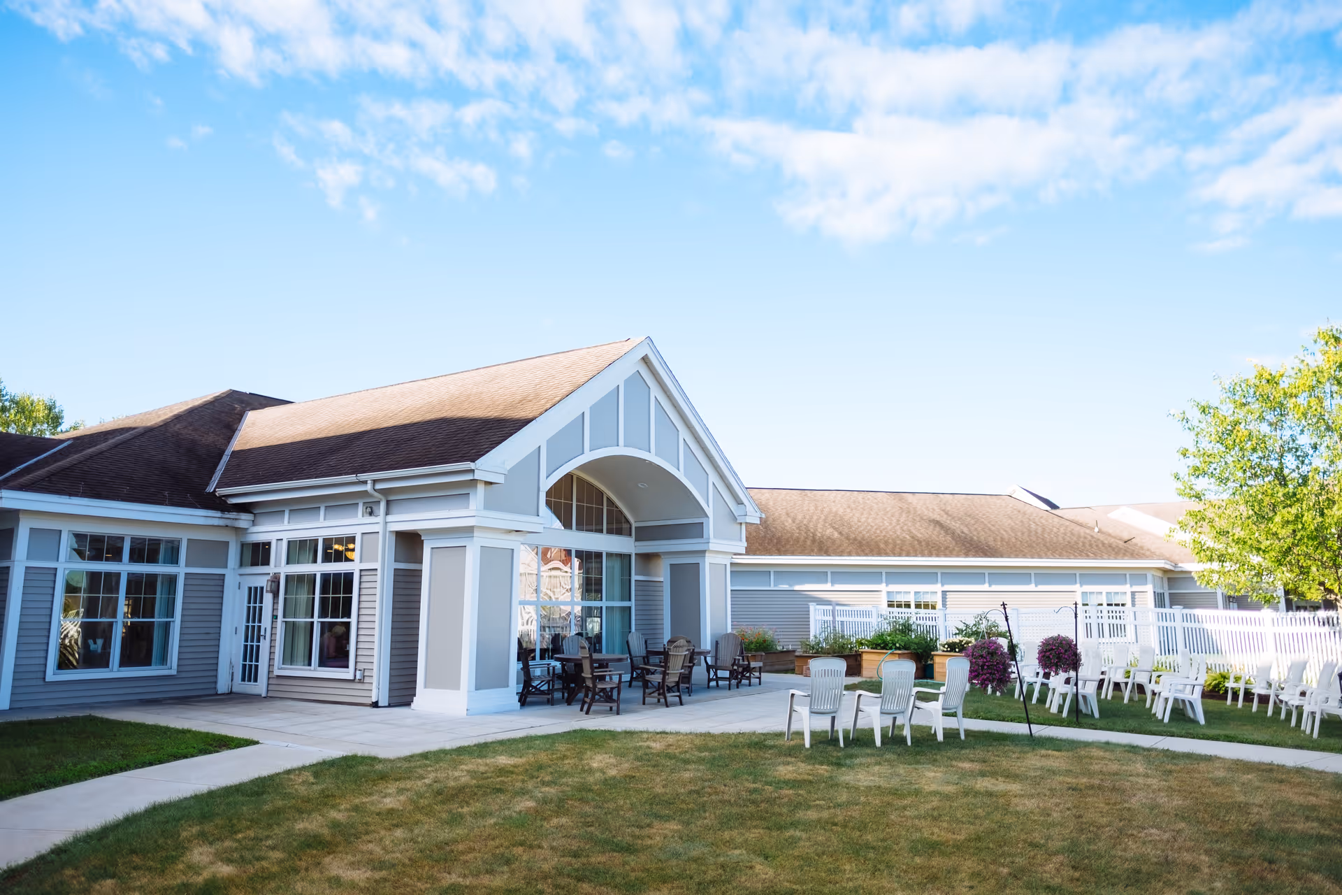 Exterior view of a senior living facility building with a light gray and white facade, large windows, and a covered patio area with outdoor seating. The foreground shows a grassy lawn with several white chairs arranged in groups, and a clear blue sky with some clouds above.