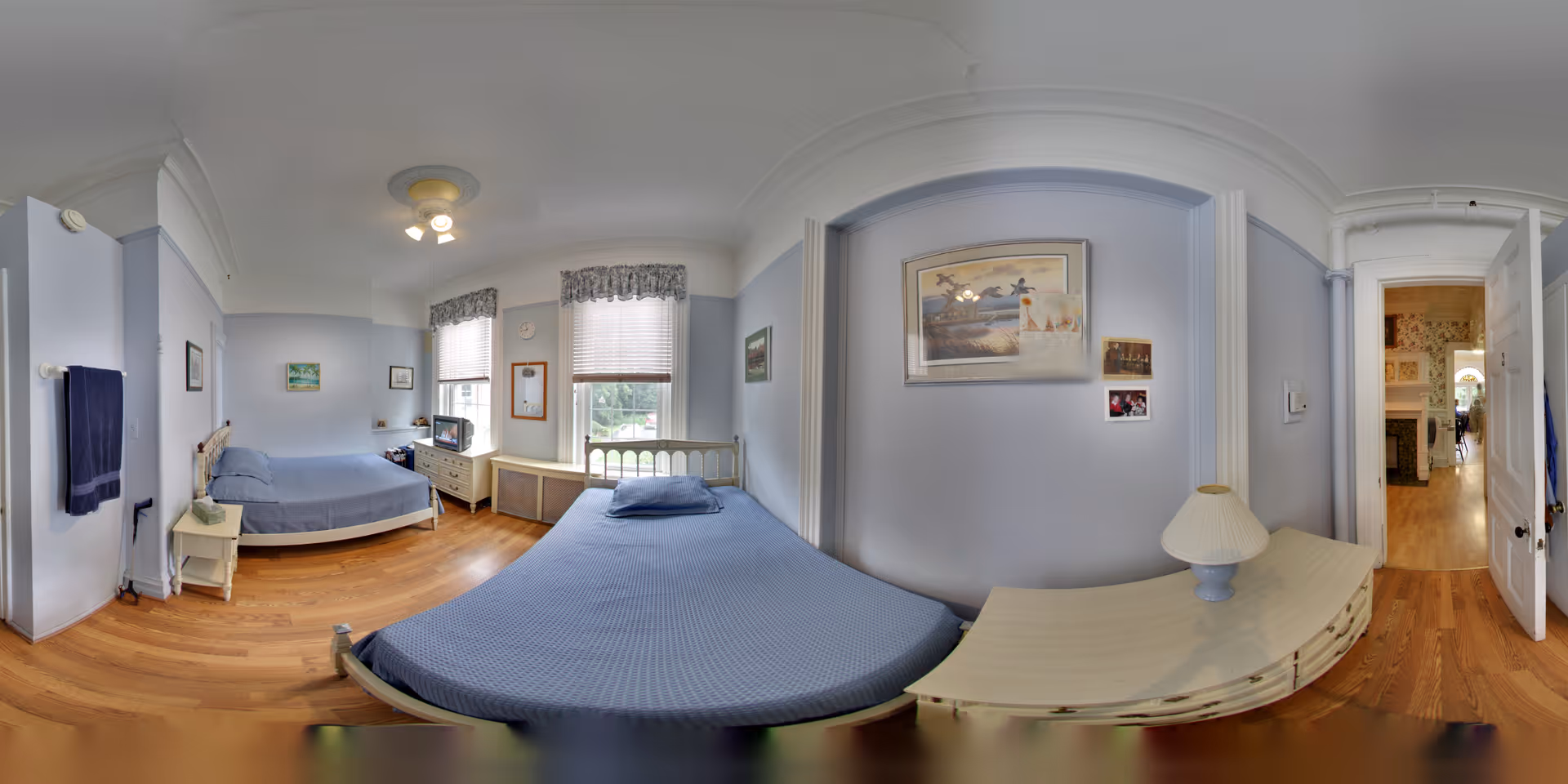 A residential care home bedroom with two beds covered in blue bedspreads. The room has light blue walls, wooden flooring, and two windows with blinds and valances. There is a small white nightstand with a lamp, a dresser with a TV, and framed pictures on the walls. The door is open, showing a hallway with wooden floors and a fireplace in the adjacent room.