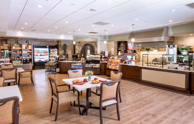 A spacious dining area with several tables and chairs, featuring a food service counter and refrigerated display cases stocked with various food items. The room has wooden flooring, pendant lighting, and shelves with additional products in the background.