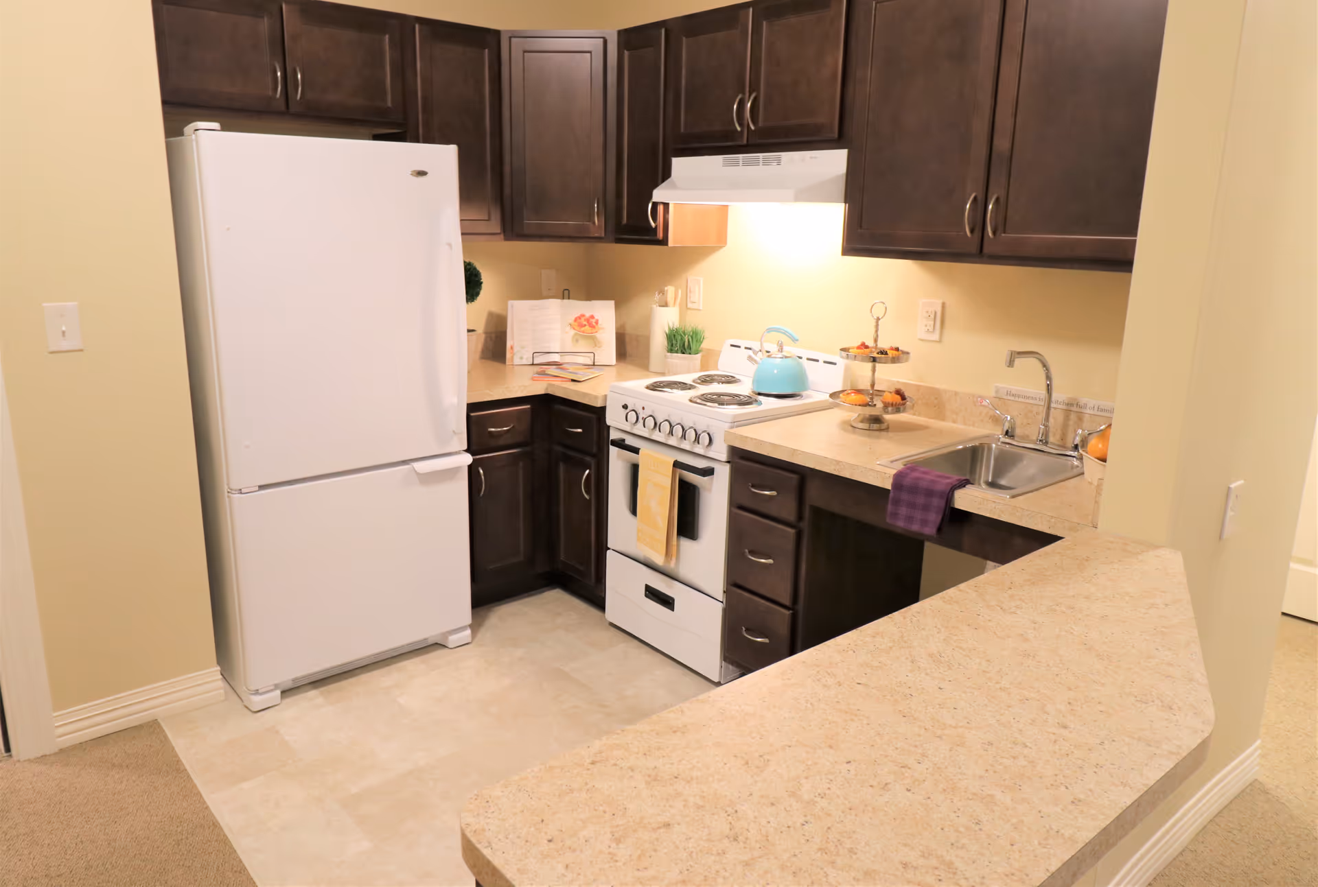 A compact kitchen with dark wooden cabinets, a white refrigerator, a white electric stove with a blue kettle on top, a sink with a purple towel hanging over the edge, and beige countertops. There are some decorative items including a two-tiered tray with pastries, a small plant, and an open cookbook on the counter.