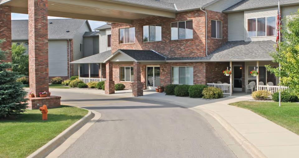 Exterior view of a senior living facility with a covered entrance supported by brick pillars, surrounded by green lawns, shrubs, and a sidewalk. The building has multiple windows and a brick facade.