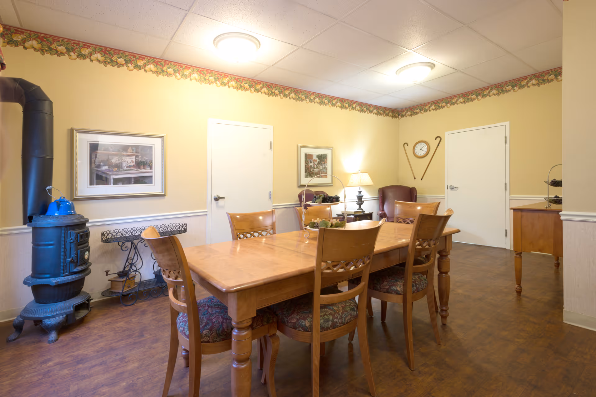 A cozy dining room with a wooden table and six chairs with patterned cushions. The room has yellow walls with a floral border near the ceiling, two closed white doors, a framed picture on the wall, a vintage black stove with a blue kettle on top, a side table with a lamp, and a clock on the wall flanked by two walking canes.