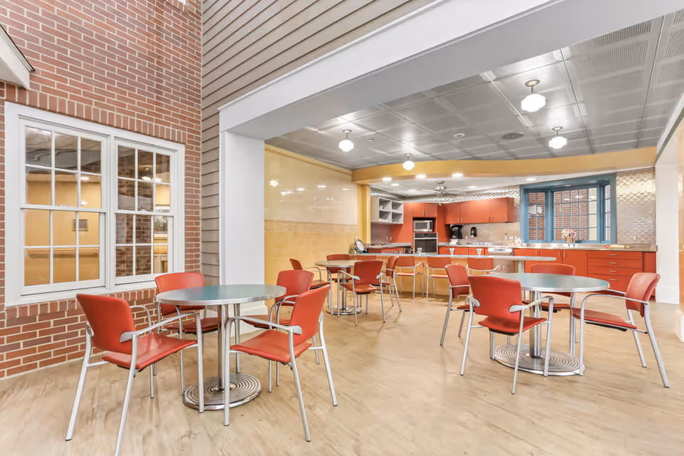 A bright and clean dining area with several round tables and red chairs. The room features a kitchen area in the background with red cabinets, a microwave, and a window. The walls have a mix of brick and beige tiles, and the ceiling has recessed lighting fixtures.