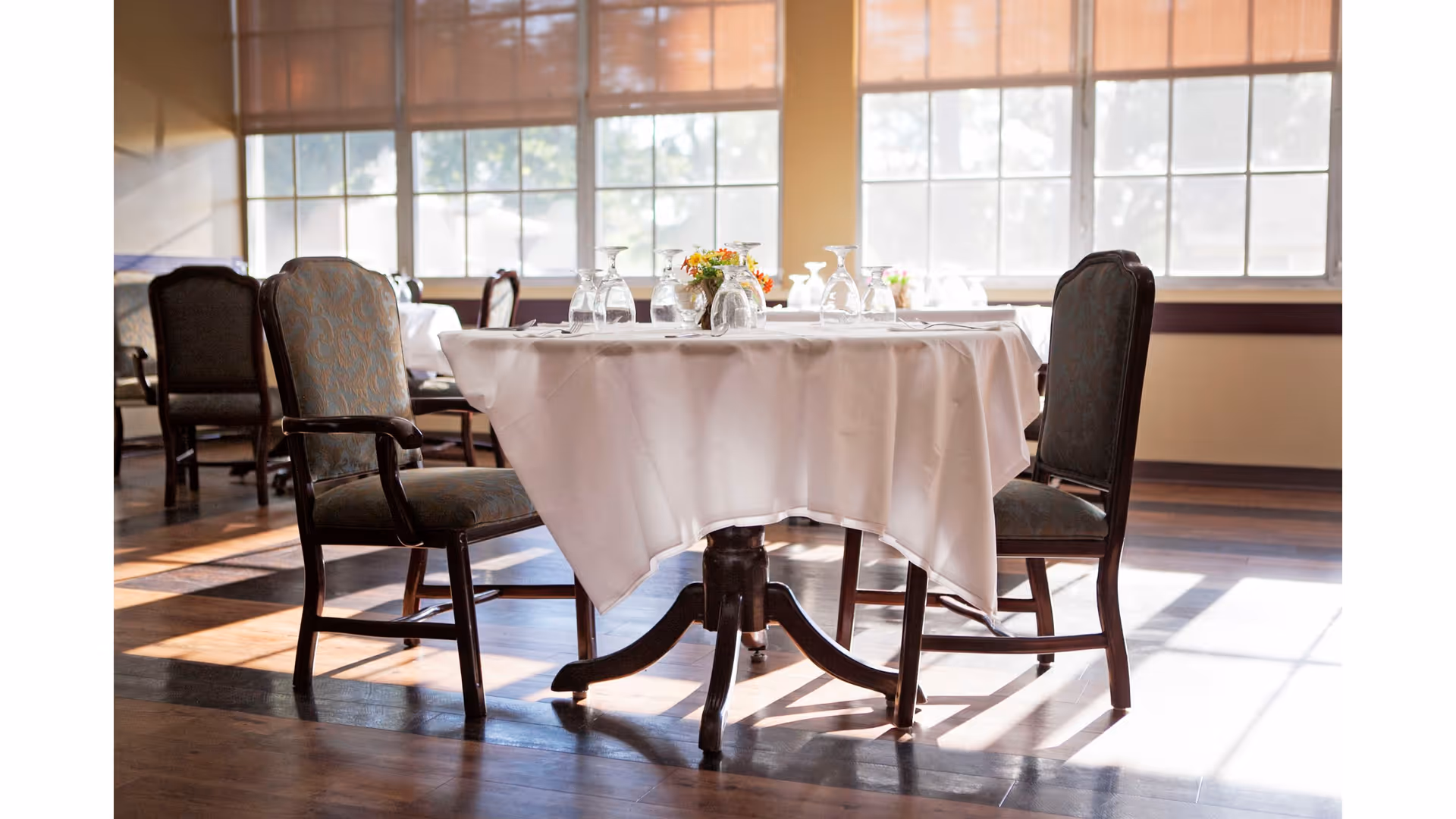 A dining table set with a white tablecloth and upside-down wine glasses in a sunlit room with large windows and wooden floors. Four upholstered chairs surround the table.