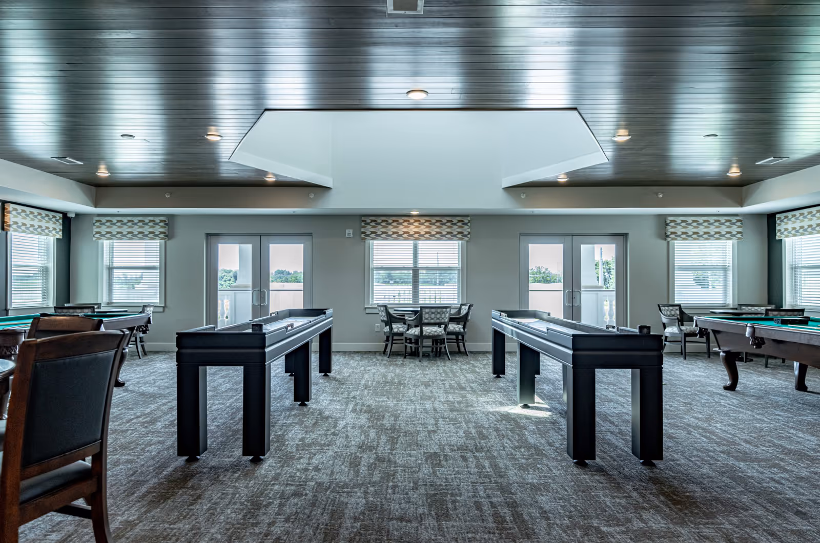 A spacious game room with two shuffleboard tables in the center, pool tables on either side, and several chairs and small tables near windows with patterned valances. The room has a modern ceiling with recessed lighting and large windows letting in natural light.