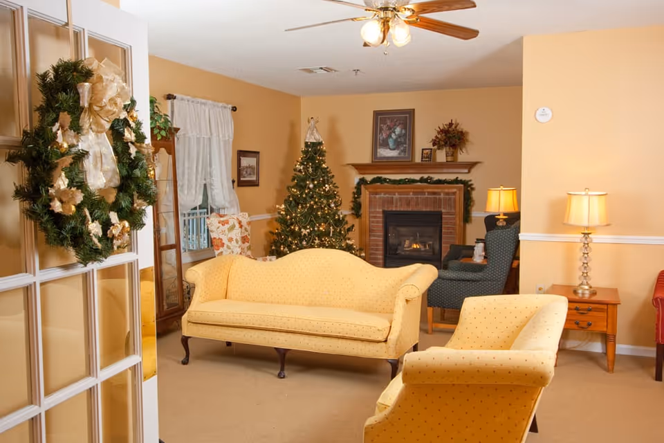 A cozy living room decorated for Christmas with a yellow sofa and armchair, a decorated Christmas tree, a brick fireplace with garland, a ceiling fan with lights, and a wreath hanging on a glass-paneled door.