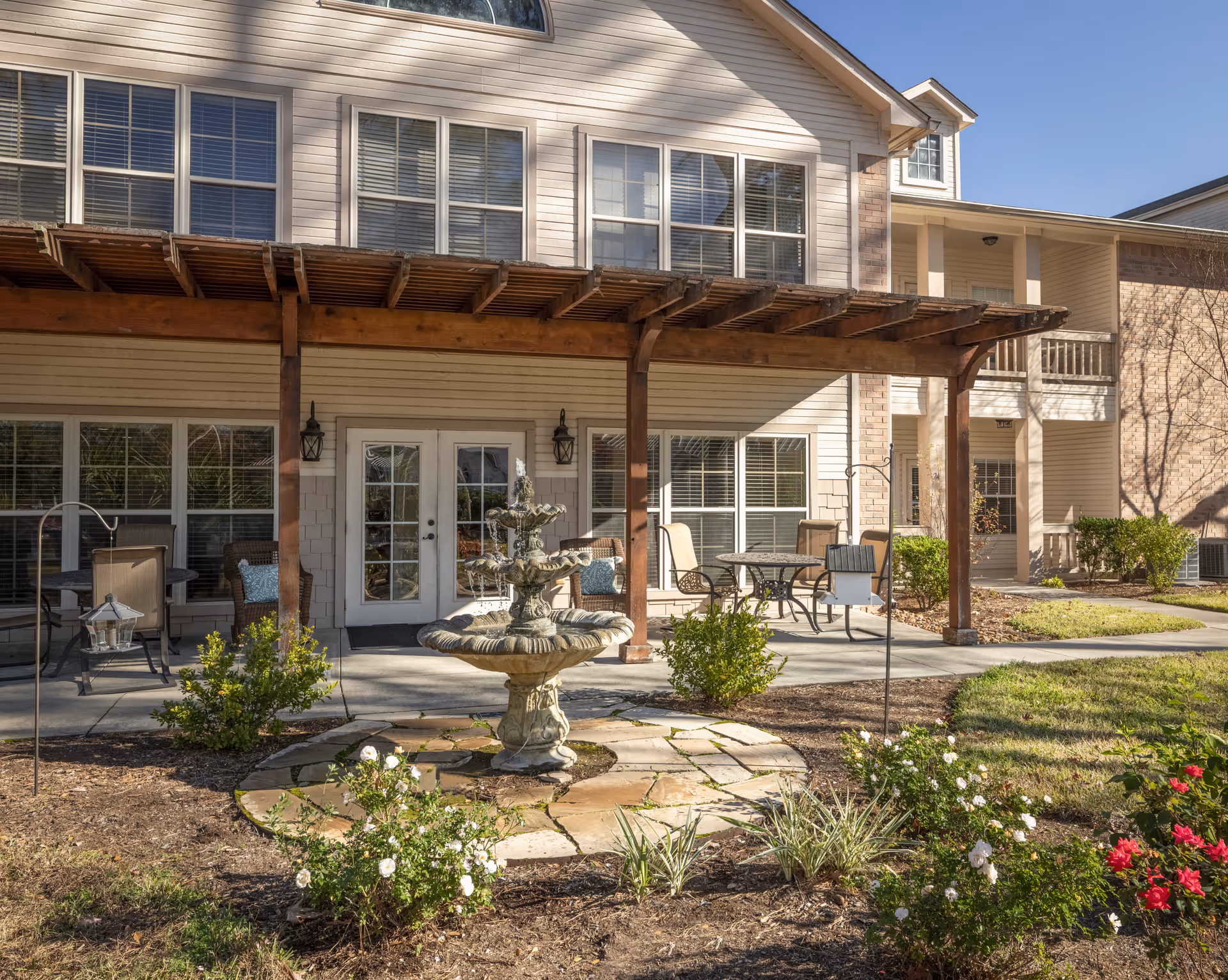 Outdoor patio area of a senior living facility with a stone fountain in the center surrounded by flower beds and shrubs. The patio has a wooden pergola overhead and several seating arrangements with tables and chairs. The building behind has large windows and a double door entrance.