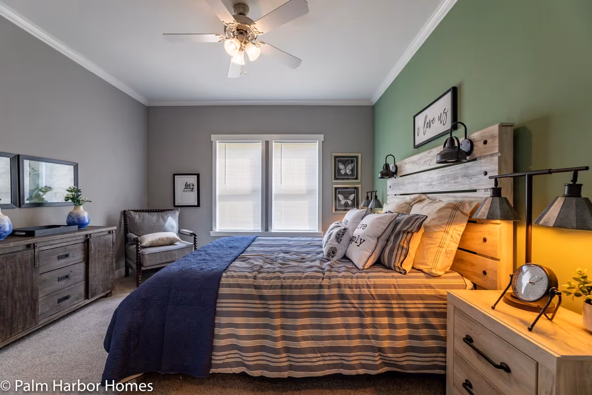 A cozy bedroom with a large bed featuring striped bedding and multiple pillows. The headboard is wooden with two wall-mounted lamps. The wall behind the bed is painted green with a framed sign that says 'i love us'. There is a wooden nightstand with a clock and a small plant, a dresser with two blue vases and a mirror above it, and a cushioned chair near two windows with blinds. The ceiling has a white fan with lights.