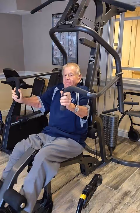 An elderly man using a weight machine for strength training in a fitness room with wooden flooring and exercise equipment in the background.