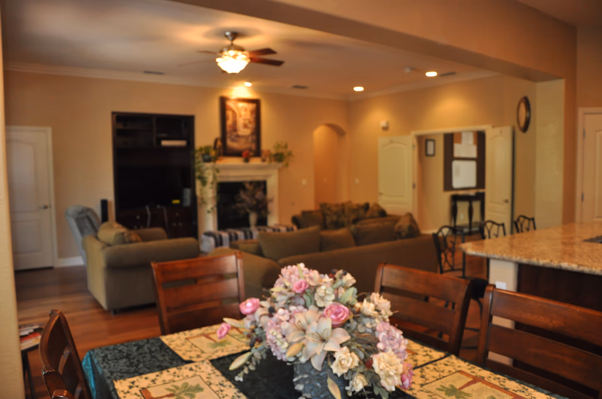 Interior view of a senior living facility showing a dining table with a floral centerpiece in the foreground, and a living room area with sofas, a fireplace, and a TV in the background. The room has warm lighting and beige walls.