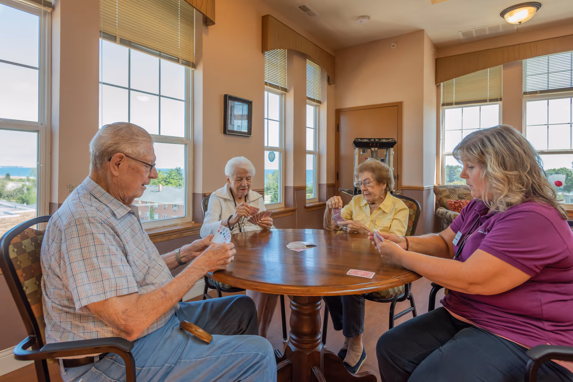 Four people sitting around a round wooden table playing cards in a bright room with large windows. Three elderly individuals and one younger woman are engaged in the game, with natural light coming through the windows behind them.
