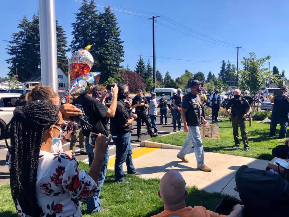 A group of people gathered outdoors on a sunny day near a building entrance, some holding balloons and taking photos.