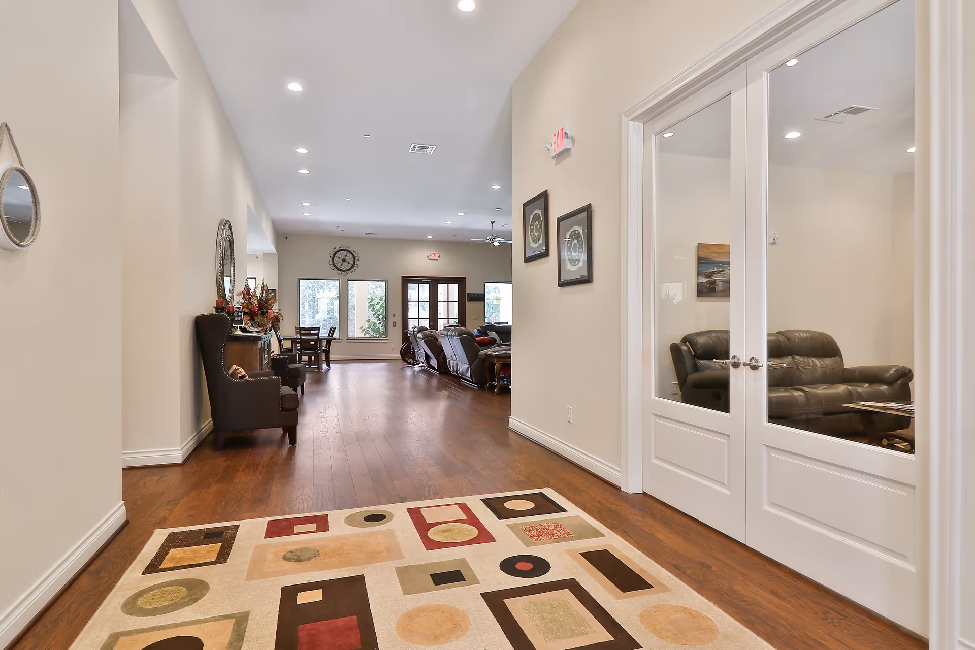 A spacious hallway in an assisted living facility with wooden flooring and a patterned area rug. On the right, there are glass double doors leading to a room with black leather sofas. On the left, there are chairs and a decorative mirror on the wall. At the end of the hallway, there is a dining area with tables and chairs, large windows, and a clock on the wall.