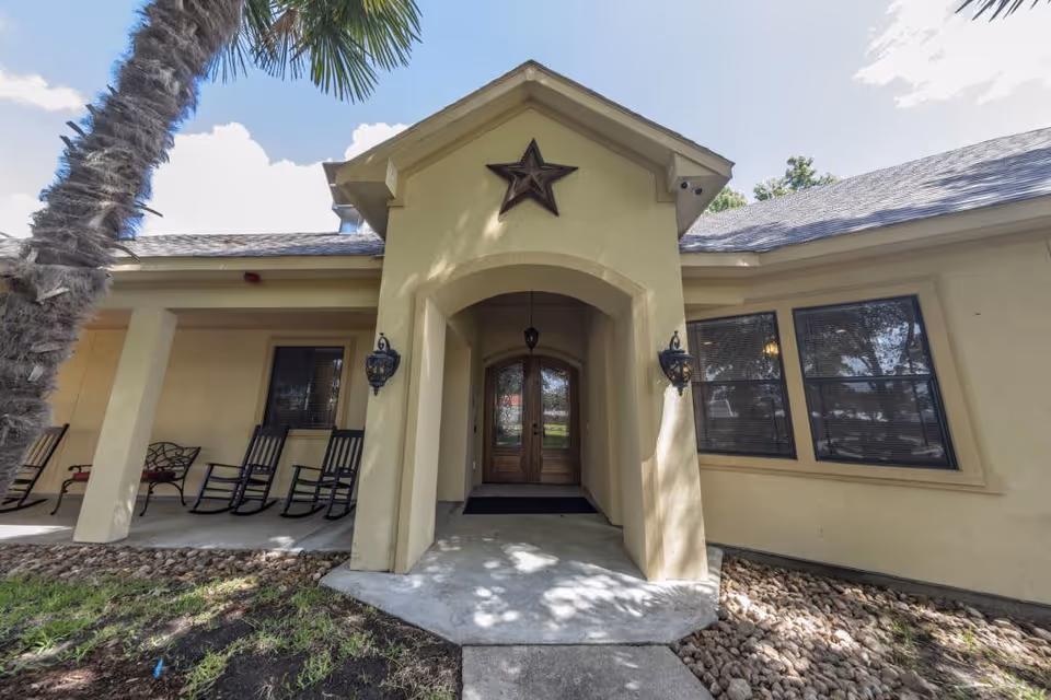 Front entrance of a senior living facility with a covered porch, two black rocking chairs, a bench, and a large star decoration above the doorway. The building is light yellow with a shingled roof and has windows with blinds.