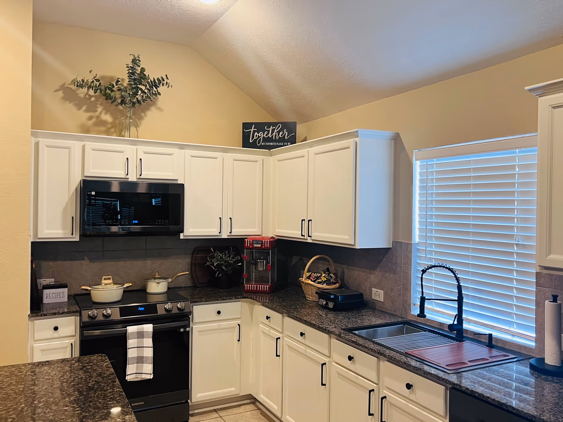 A modern kitchen with white cabinets, a black stove with a white pot and pan on it, a microwave above the stove, a black countertop with a red popcorn machine, a basket of snacks, a sink with a black faucet, a window with white blinds, and a decorative sign that says 'together our favorite place to be' on top of the cabinets.