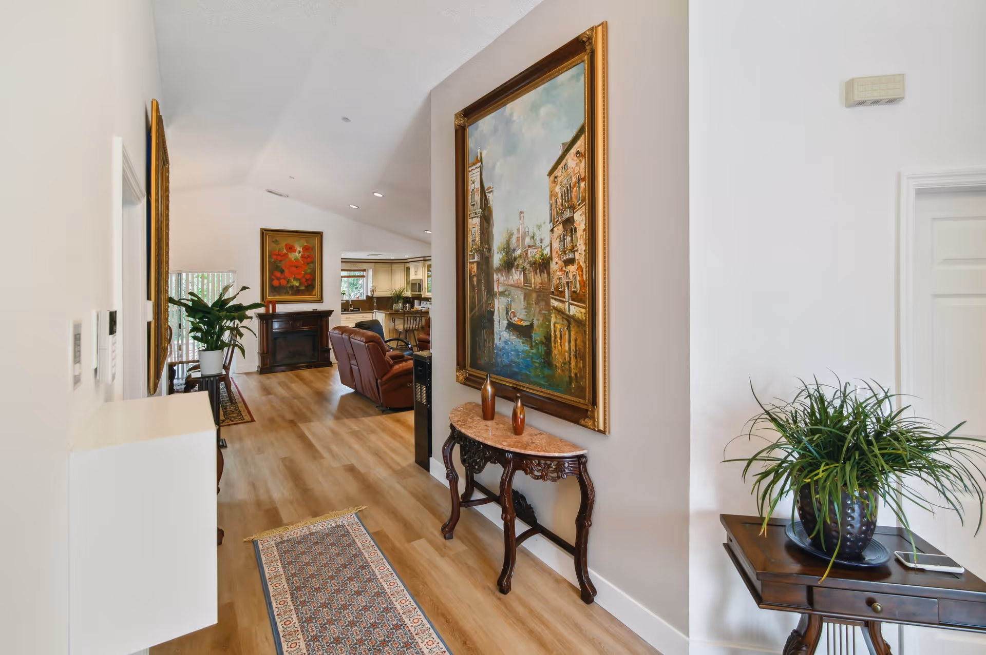 Interior view of a senior living facility hallway with wooden flooring, a decorative rug, a small ornate table with two vases, a large framed painting on the wall, and a side table with a potted plant and a smartphone. The hallway leads to a living area with a brown leather recliner, a fireplace, another painting, and a kitchen area in the background.