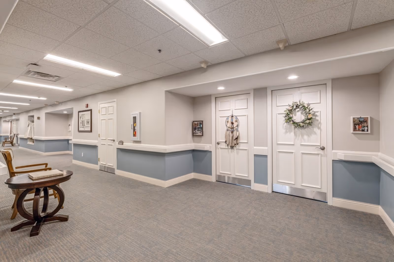 A clean and well-lit hallway in a senior living facility with light gray walls, blue wainscoting, and carpeted floors. There are two white doors decorated with a dreamcatcher and a wreath, a small wooden table with a book on it, and a wooden chair. The ceiling has recessed lighting and a drop ceiling with fluorescent lights.