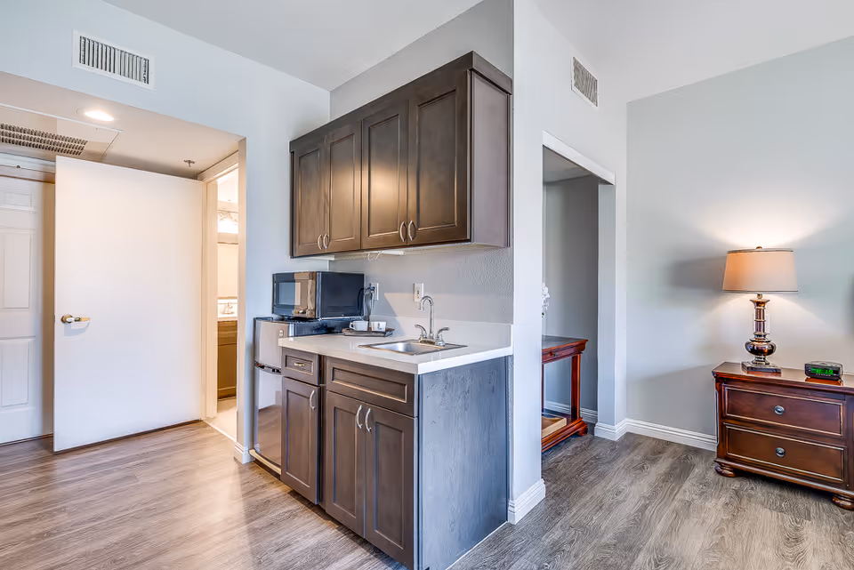 Kitchenette with dark upper and lower cabinets, a sink, microwave and mini-fridge next to a bedside table and lamp.