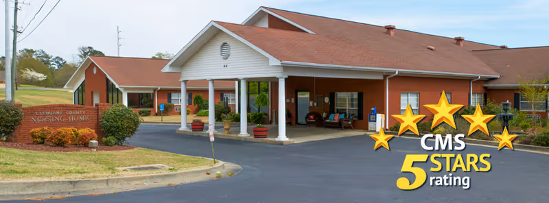 Exterior view of Cleburne County Nursing Home showing a single-story brick building with a covered entrance supported by white columns, surrounded by landscaped bushes and a paved driveway.