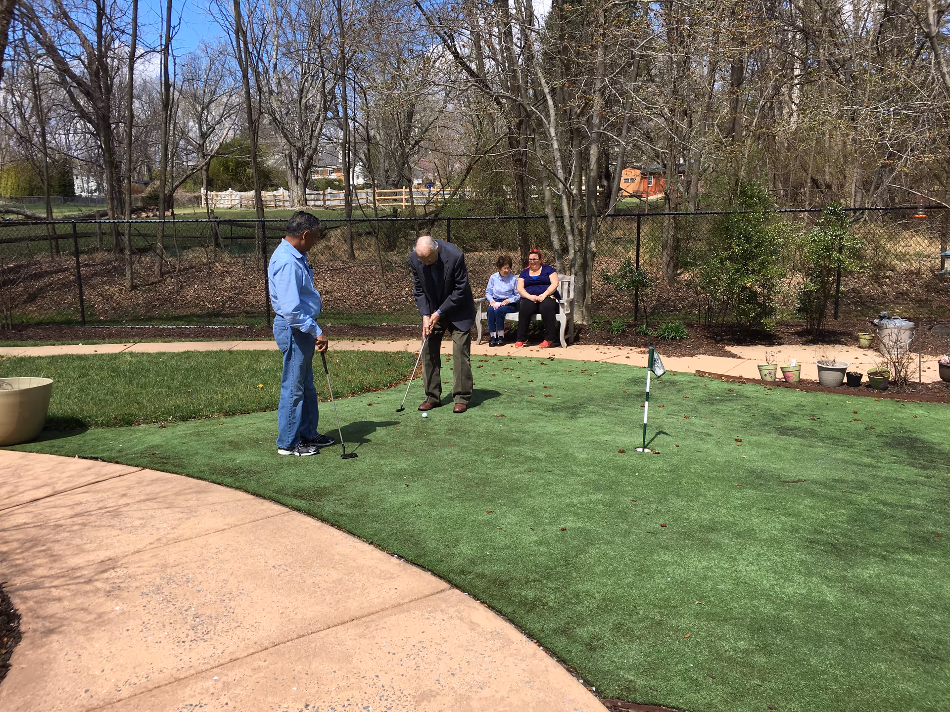 Two men practice putting on a small outdoor putting green while two women sit on a bench nearby under trees.