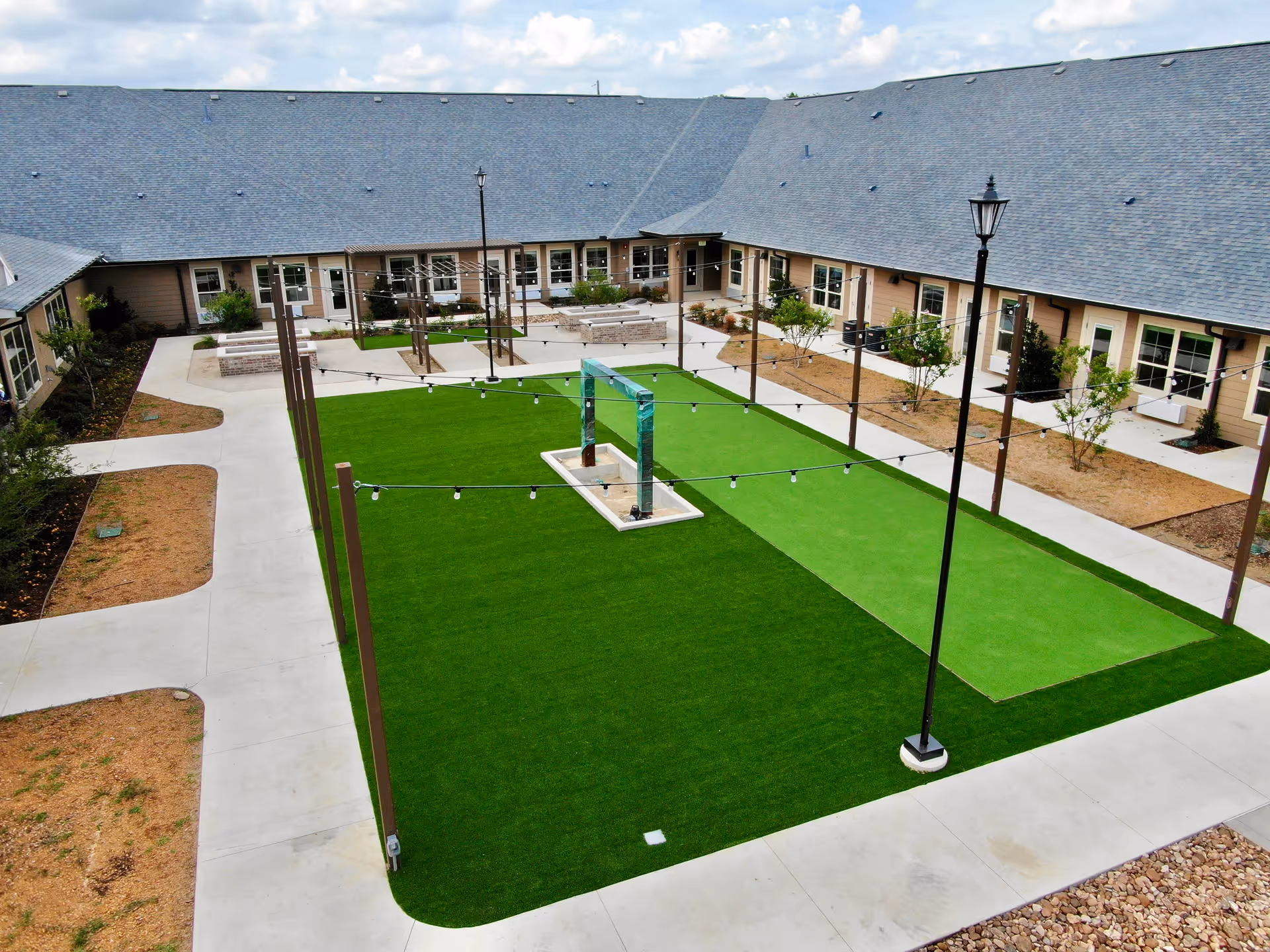 A courtyard area in a senior living facility with a large green artificial turf lawn, string lights hanging overhead, a central decorative structure, surrounded by a concrete walkway and a building with multiple windows and doors.