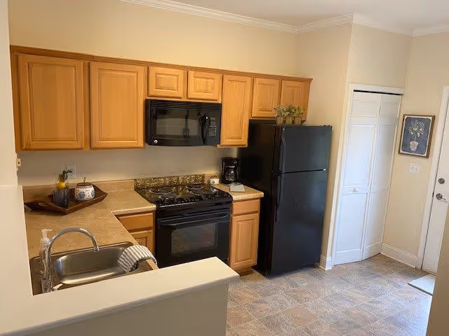A compact kitchen with wooden cabinets, a black microwave above a black stove, a black refrigerator, a stainless steel sink with a faucet, and a countertop with a few decorative items. The floor is tiled, and there is a white door and a closet with white louvered doors on the right side.