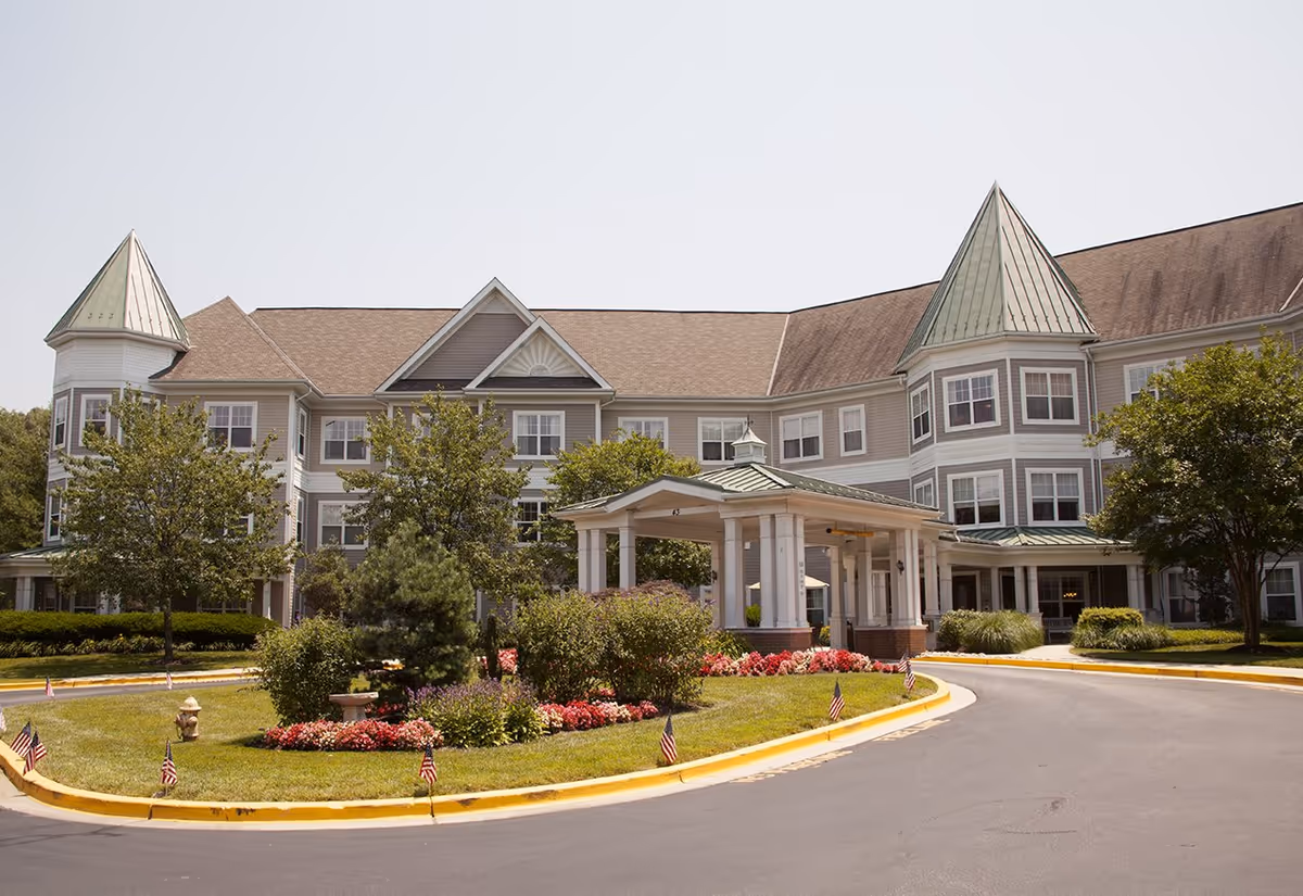 Exterior view of a large senior living facility building with two green-roofed towers, multiple windows, and a covered entrance. The driveway curves around a landscaped area with flowers, bushes, small trees, and small American flags. The sky is clear.