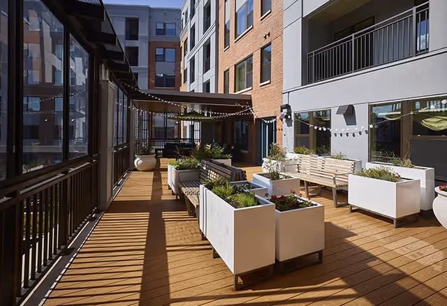 Sunny outdoor courtyard terrace with wooden decking, benches, planter boxes and string lights between apartment buildings.