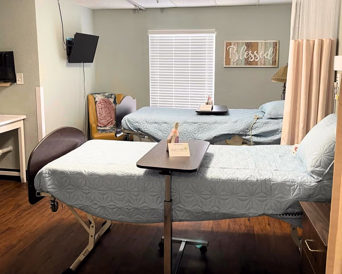 Shared patient room with two hospital beds dressed in light blue quilts, overbed tables, a wall-mounted TV, a recliner, and a 'Blessed' sign above the window.