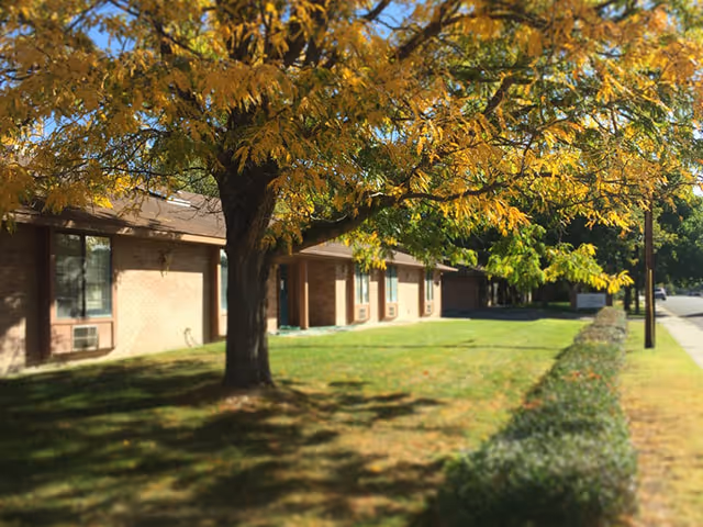 Exterior view of a single-story brick senior care building with trees, lawn, and a sidewalk.