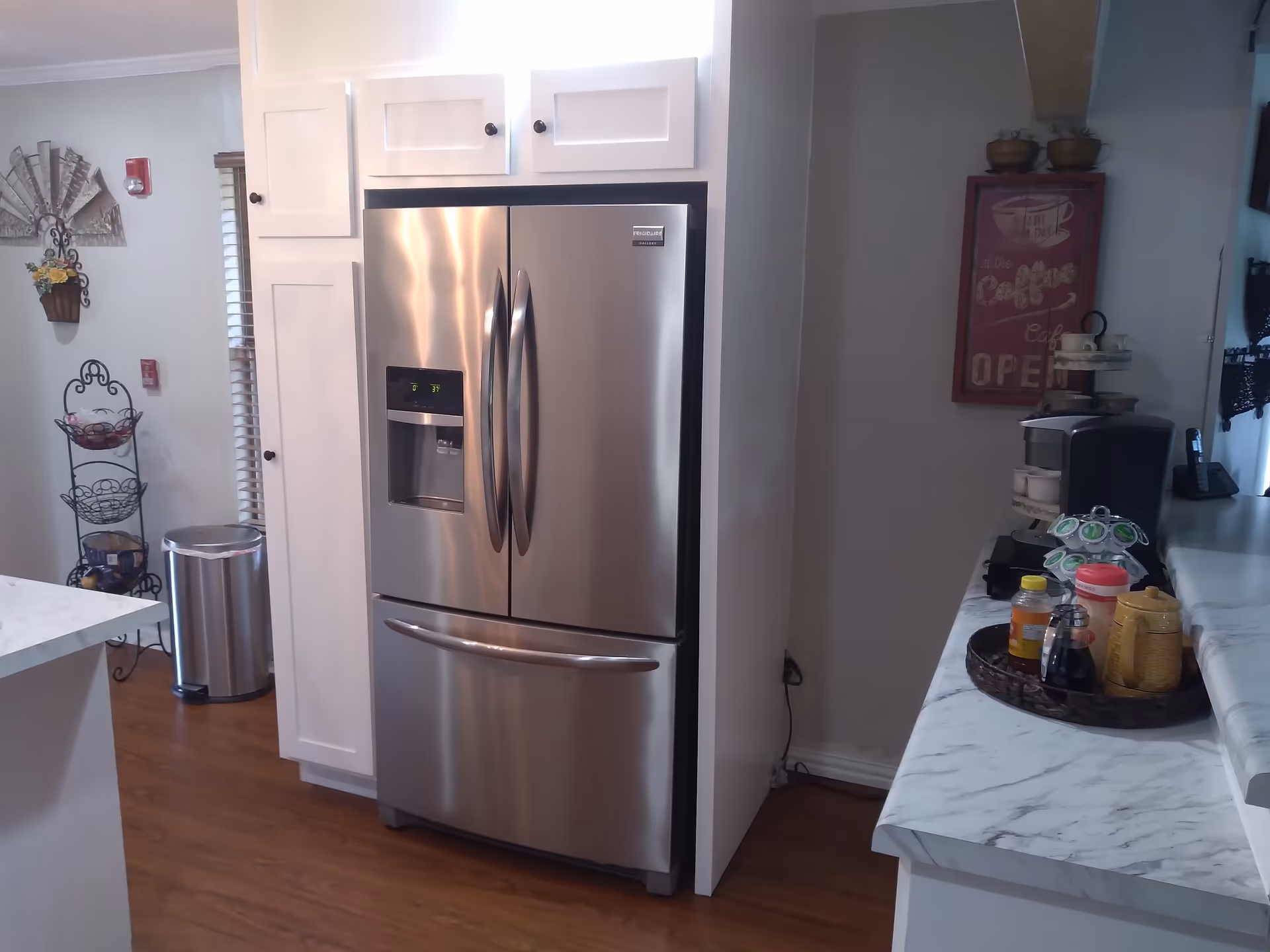 A modern kitchen area featuring a stainless steel French door refrigerator with a bottom freezer drawer, white cabinetry, a marble countertop with various kitchen items including a coffee maker, condiments, and a rotating coffee pod holder. The floor is wooden, and there is a decorative metal rack with baskets and a trash can in the background.