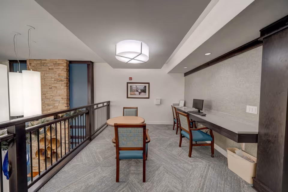 Carpeted interior common area with a round table and chairs, a long wall-mounted desk with computers, and a railing overlooking an atrium.