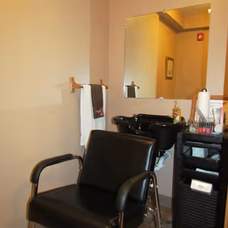 Interior of a salon area with a black salon chair in front of a black hair washing sink. A large mirror is mounted on the wall above the sink. A towel rack with white and dark towels is visible on the wall. A small cart with various hair care products and supplies is next to the sink.